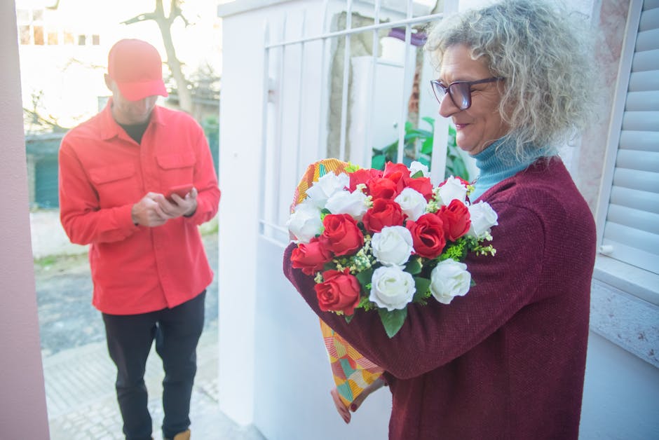A woman with curly gray hair and glasses is standing beside a doorway, holding a colorful bouquet of fresh red and white roses wrapped in decorative paper. She is wearing a blue turtleneck and a maroo