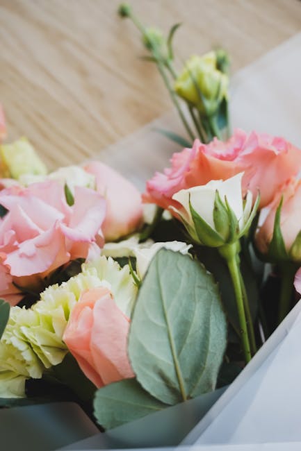 Close-up of a floral gift featuring a bouquet of fresh lisianthus flowers in pastel shades of pink, cream, and pale yellow, with green buds and stems, wrapped in white paper with a smooth, matte textu