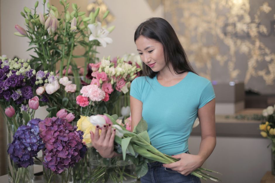 A young woman with long dark hair wearing a light blue t-shirt is standing in a floral shop, carefully holding a bouquet of light yellow tulips with green stems and leaves. She is surrounded by an arr