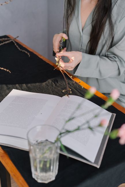 A woman wearing a grey blazer is arranging a bouquet of fresh flowers, with a pair of scissors in hand, at a wooden table. The flowers include pink and green blooms with slender stems, and the bouquet