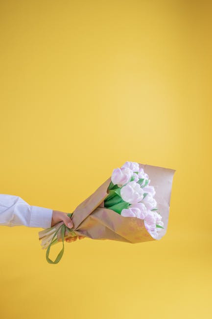 A person is holding a bouquet of pale pink tulips wrapped in light brown paper with a green ribbon, against a bright yellow background. The tulips have smooth, rounded petals with fresh green stems vi