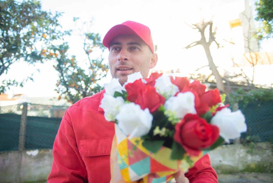 A male flower delivery courier wearing a red cap and matching red jacket holding a vibrant bouquet of fresh red and white roses, with green foliage interspersed, wrapped in colorful patterned paper. T