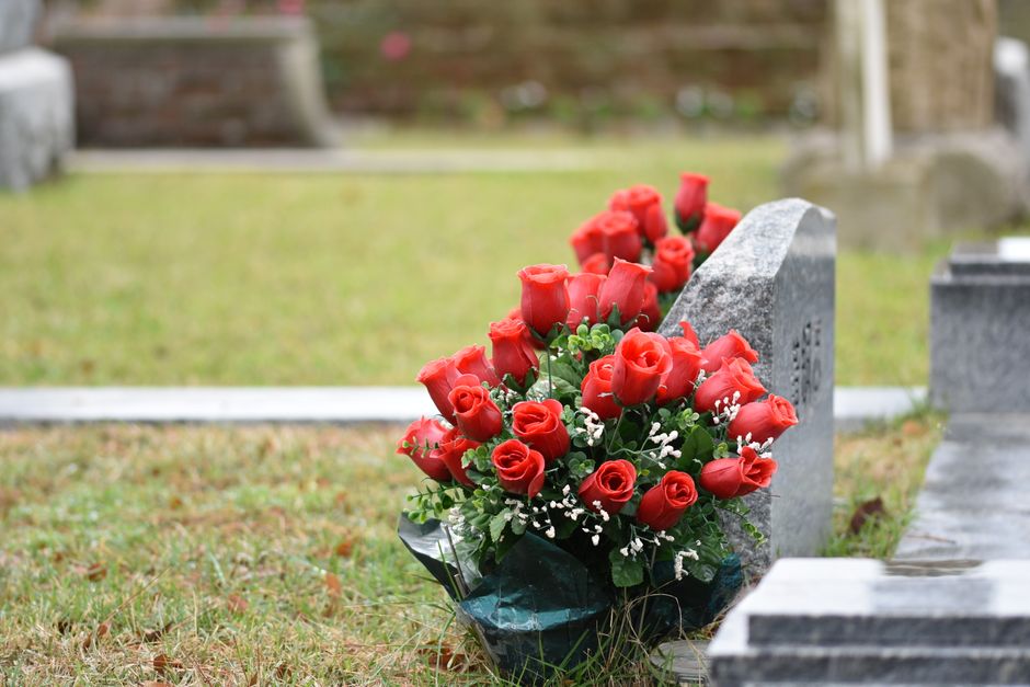 A bouquet of fresh red roses arranged in a rounded, floral style with green foliage and small white filler flowers, wrapped in dark green paper. The bouquet is placed at the base of a grey headstone i