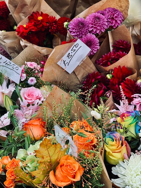 A vibrant assortment of fresh flowers including bright red dahlias and pink chrysanthemums arranged in brown paper wrapping, with some bouquets featuring pastel pink roses, orange roses, and multicolo