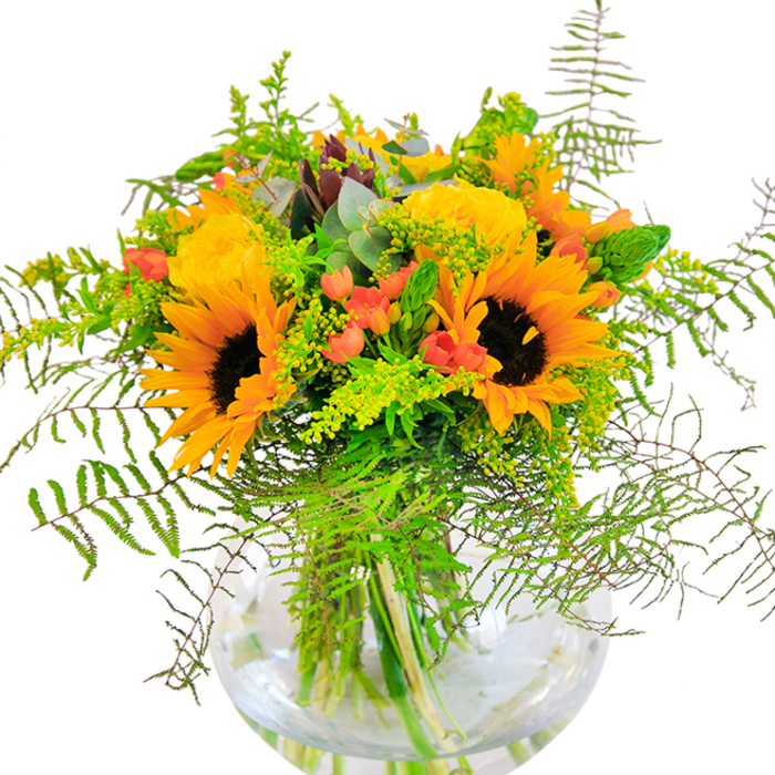Sun-infused and expressive, this bouquet from Kensal Green Florist presents a dazzling collection of yellow sunflowers and garden roses, illuminating any room much like a golden afternoon over Kensal Green's leafy streets. The sunflowers' resonant yellow petals and velvety brown cores draw the gaze, while rich yellow roses, with their compact, furled petals, lend an aristocratic poise to the arrangement. Glossy red Hypericum berries, scattered throughout like ruby droplets, provide lively contrast and a dash of opulence. Pale green and golden filler stems, such as solidago, add a buoyant texture, reminiscent of the abundant greenery in local neighbourhood gardens. The generous addition of fern fronds and blue-green eucalyptus leaves introduces softness and depth, with every element shaped to evoke the outdoors and open air. This lively interplay is contained in a clear, fishbowl-style glass vase, offering a glimpse of water-nourished stems and bringing together modern clarity and classic floral artistry. Seen up-close, the mix of airy foliage, bright blooms, and wild forms creates a sensory tapestry that whispers of summer, companionship, and celebration-ideal for birthdays, anniversaries, or simply gifting a flourish of happiness in Kensal Green. The bouquet reflects an expert's appreciation for balance, freshness, and the particular joy of local flower gifting.
